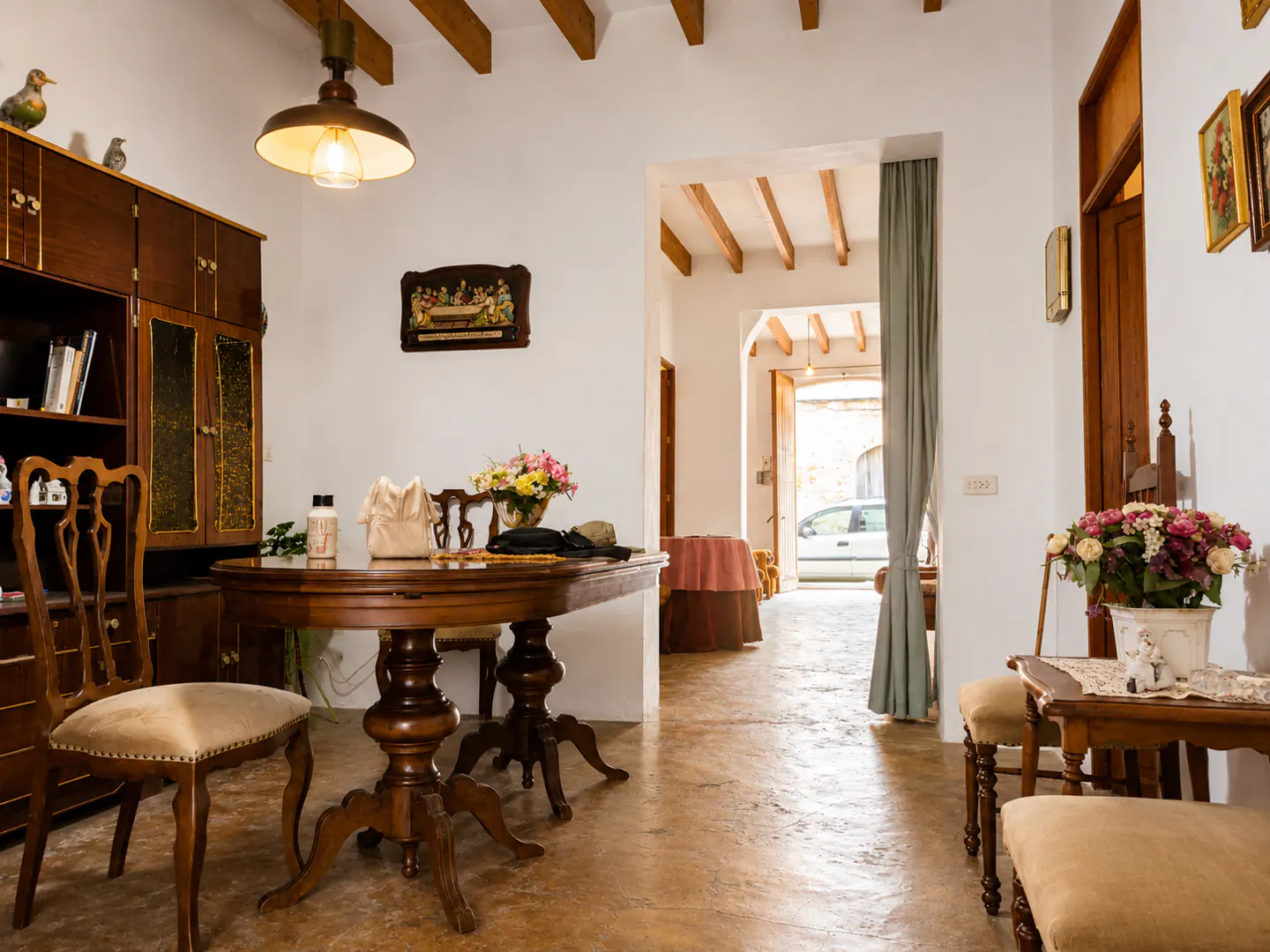 A bright living room with a wooden table, chairs, and cabinet. A doorway leads to another room with a view of a car outside.