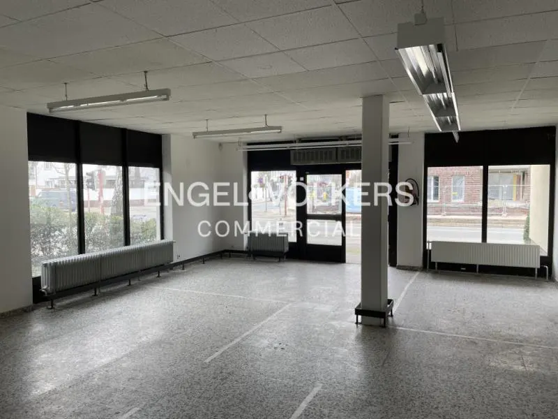 Empty commercial space with gray terrazzo floors, white walls, and black-framed windows. Radiators line the walls. Fluorescent lights illuminate the room.