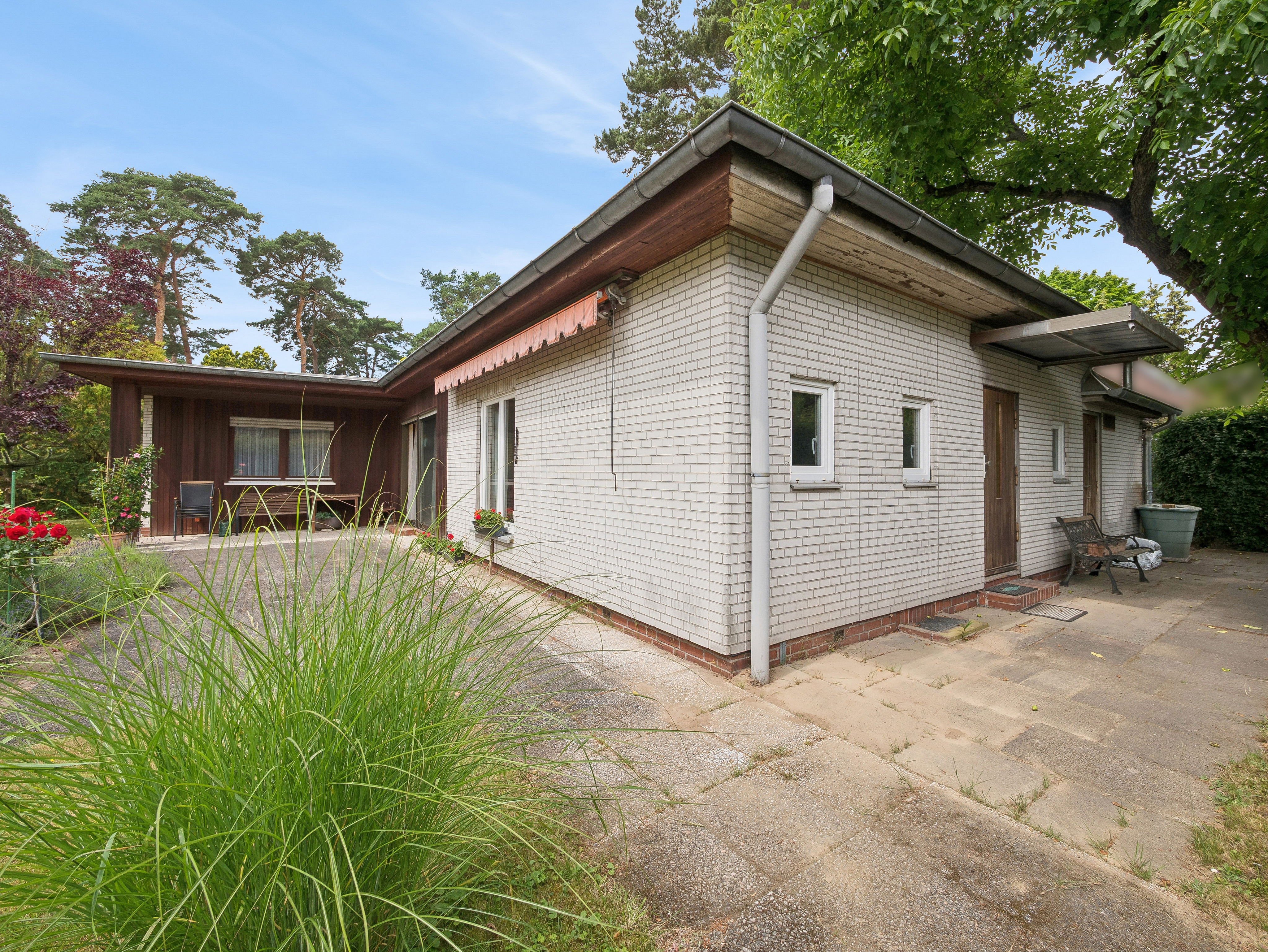 Single-story white brick house with a flat roof, wooden accents, and a paved patio area surrounded by trees and greenery.