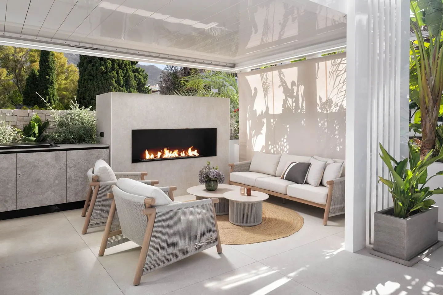 Outdoor living space with a fireplace, sofa, chairs, and coffee table on a patio with plants and trees in the background.