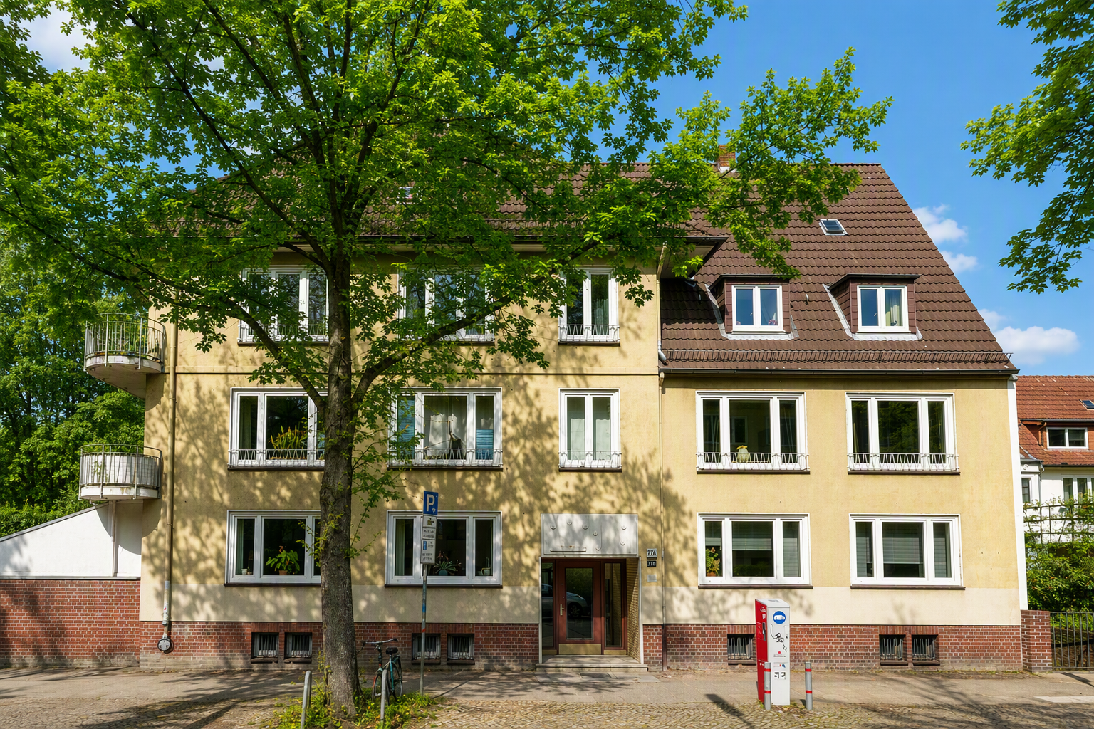 A three-story yellow apartment building with a brown tiled roof, white-framed windows, and a large tree in the foreground.