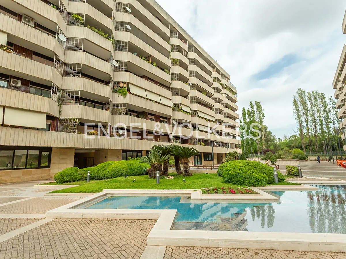 Exterior view of a beige apartment building with balconies, a pool, and green landscaping.