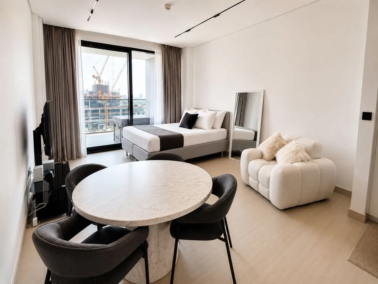 Modern studio apartment with a marble dining table, grey chairs, a bed, and a white armchair, overlooking a construction site.