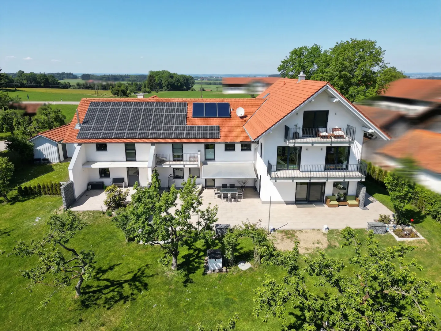 Aerial view of a white two-story house with a red tile roof and solar panels, surrounded by green lawn and trees.