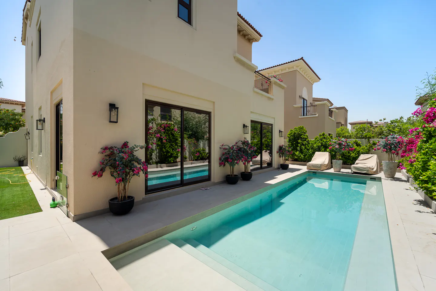 Backyard view of a two-story beige house with a blue swimming pool, potted plants, and covered lounge chairs.