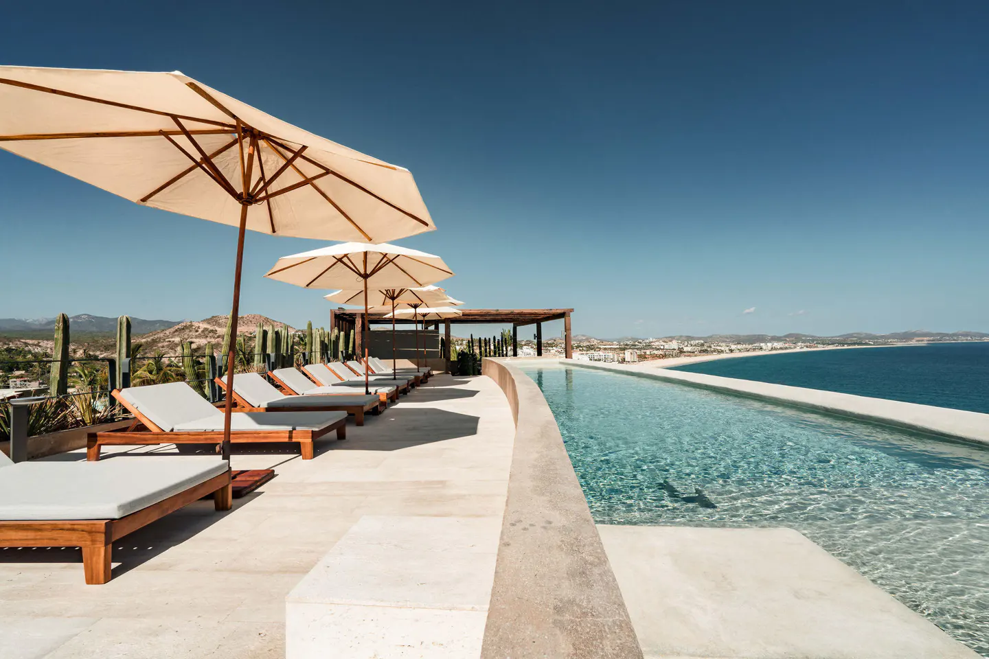 Rooftop pool with lounge chairs and umbrellas overlooking the ocean. Blue sky, mountains, and city in the background.