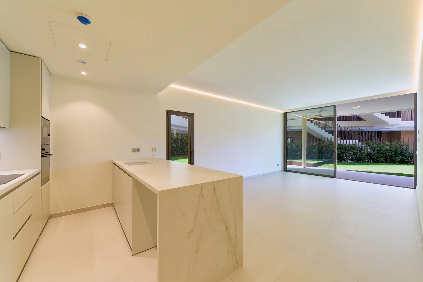 Bright, modern kitchen with white cabinets, marble island, and sliding glass doors to a green lawn and building with stairs.