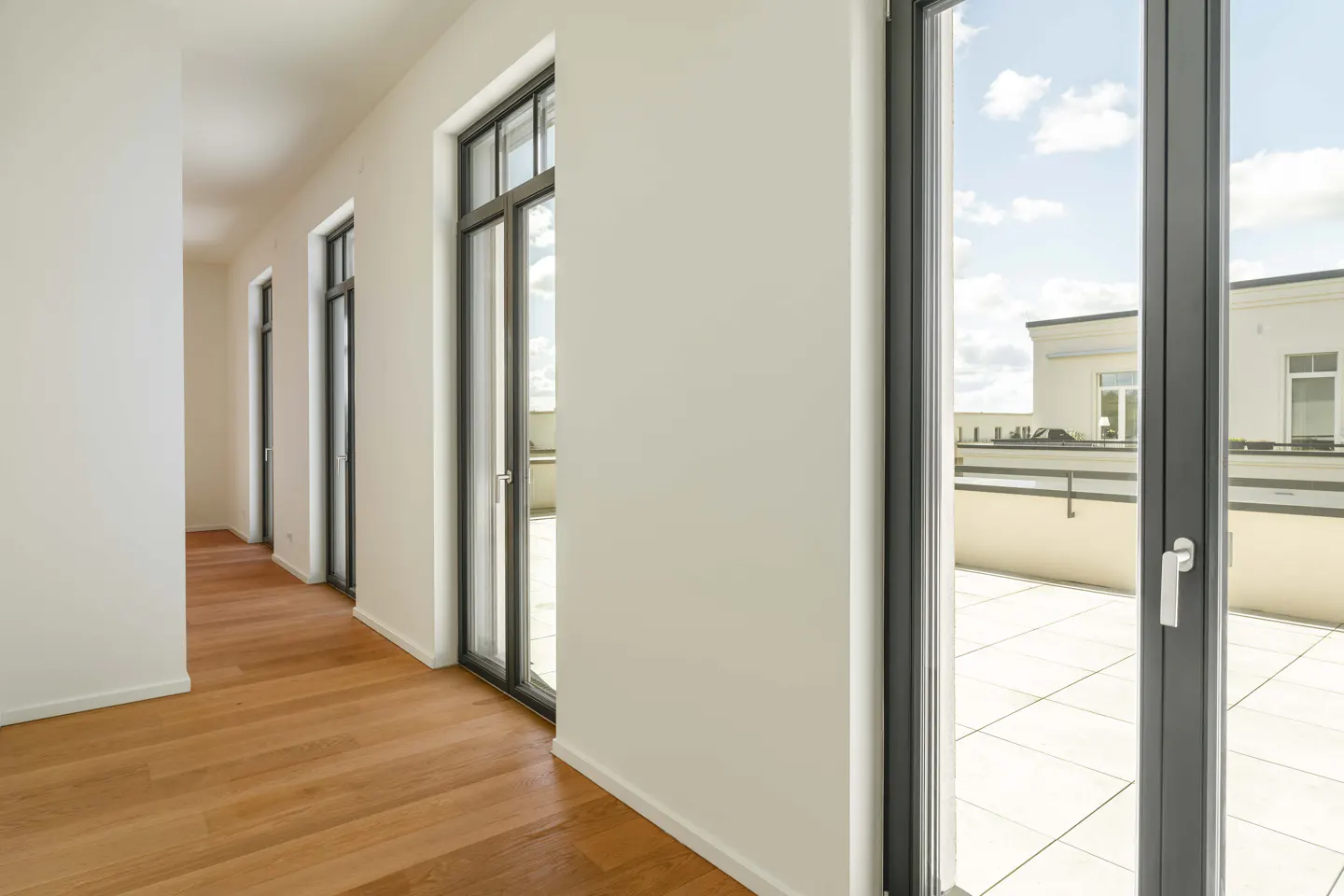 A bright hallway with wood floors and white walls leads to a balcony through black-framed glass doors.