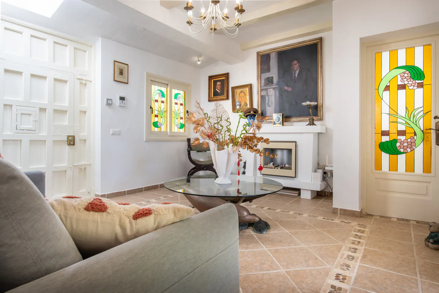 Living room with a gray sofa, glass table, and white fireplace. Portraits hang above the fireplace. A stained glass door and window add color.
