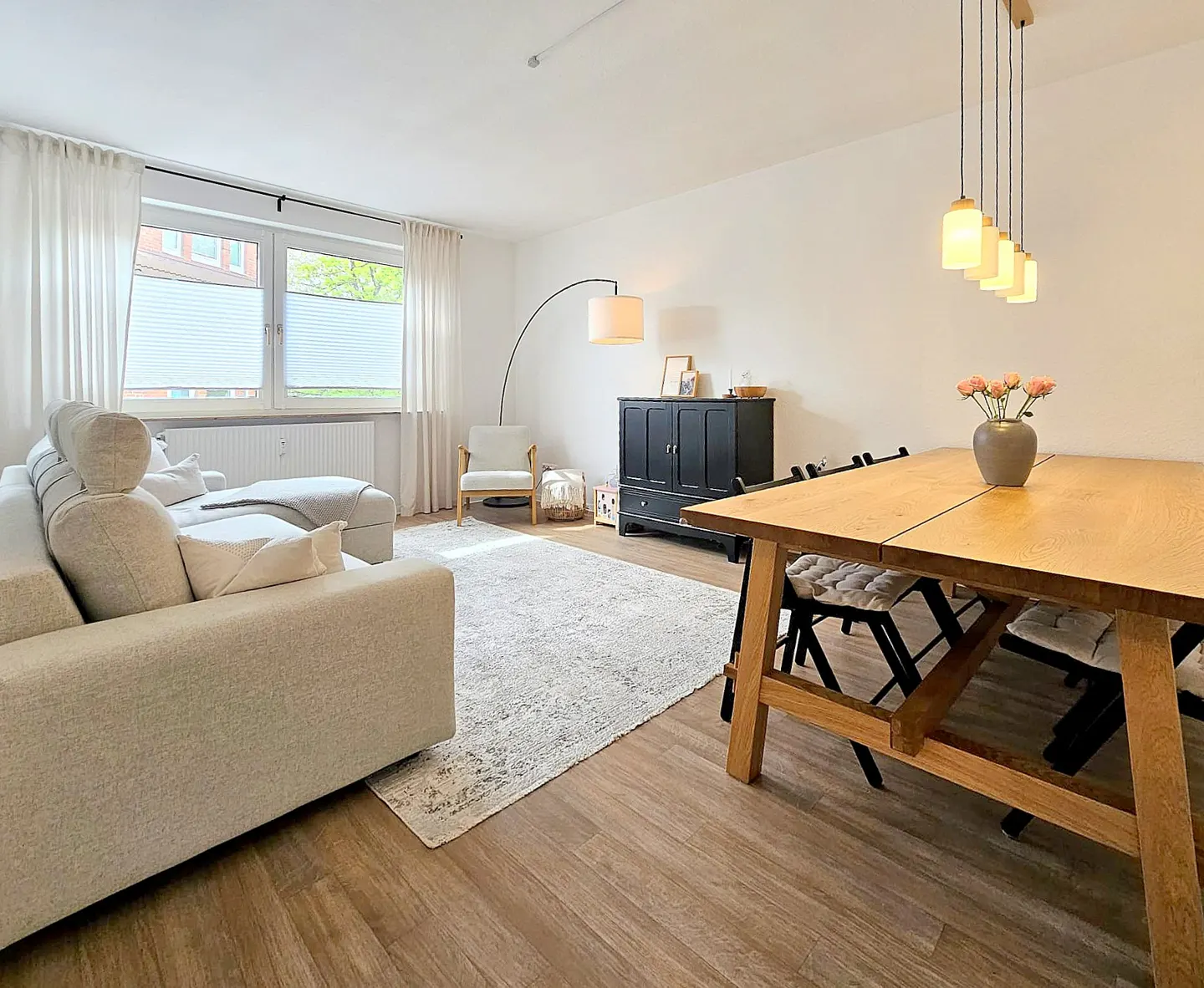 Bright living room with a beige sofa, wooden table with chairs, and a black cabinet on a light-colored rug. Natural light streams through the window.
