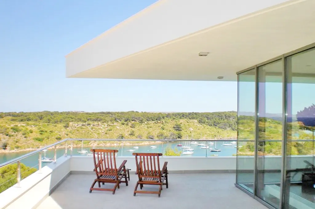 Balcony view: Two wooden chairs face a bay dotted with sailboats, framed by green hills under a clear blue sky. Modern white building with glass walls.