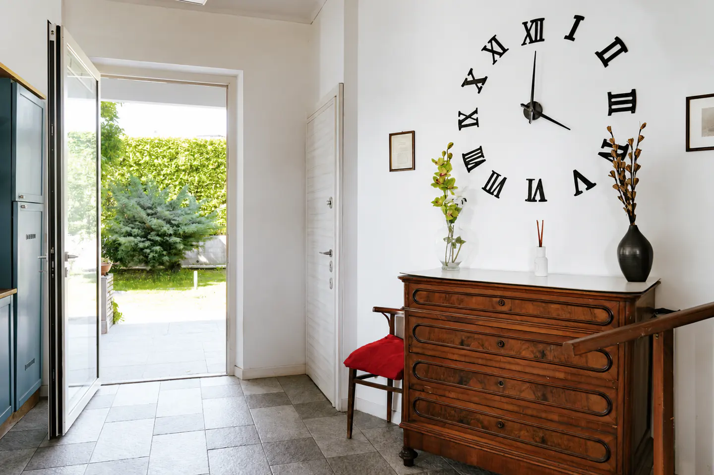 Hallway with open door to garden. A wooden dresser sits beneath a large Roman numeral clock on a white wall.