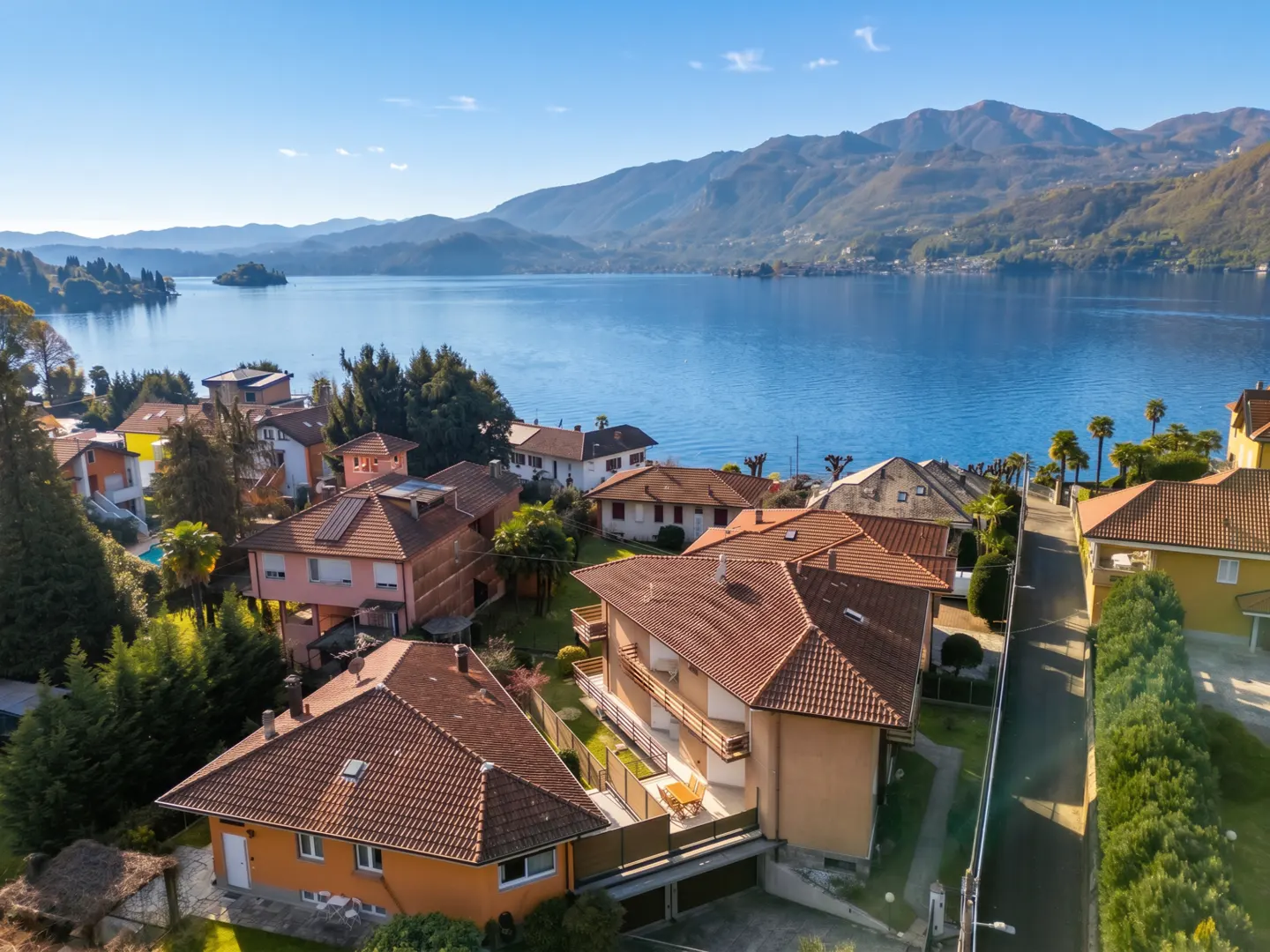Aerial view of houses with red tile roofs near a blue lake and mountains under a clear blue sky.