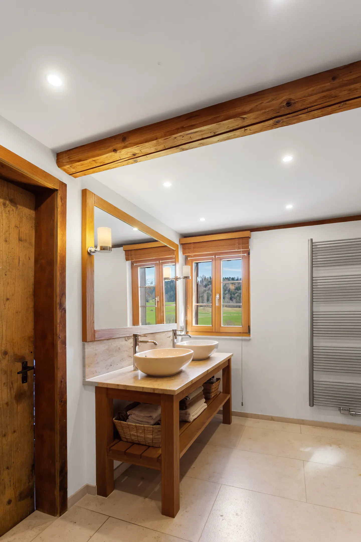 Modern bathroom with a wooden double vanity, two vessel sinks, a large mirror, and a view of green fields through windows.