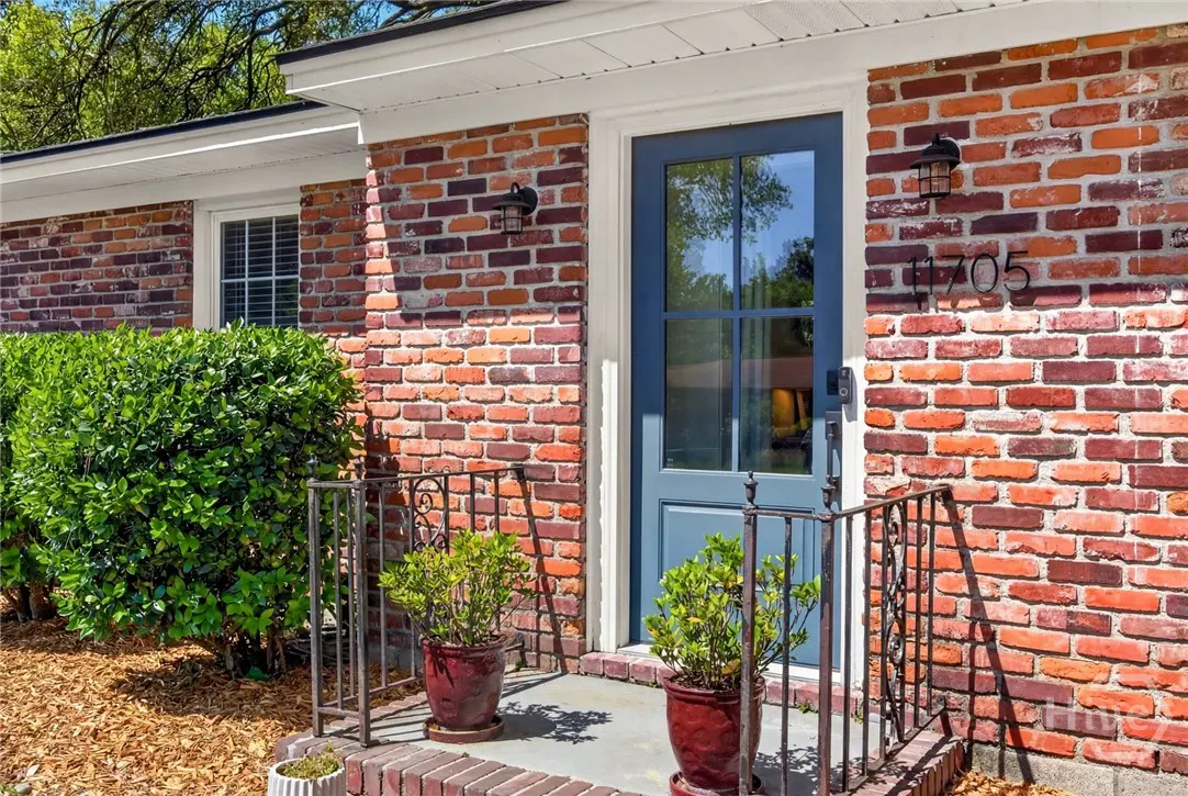 The front of a brick house with a blue door, black sconce, and the number 1705. Two potted plants flank the door.