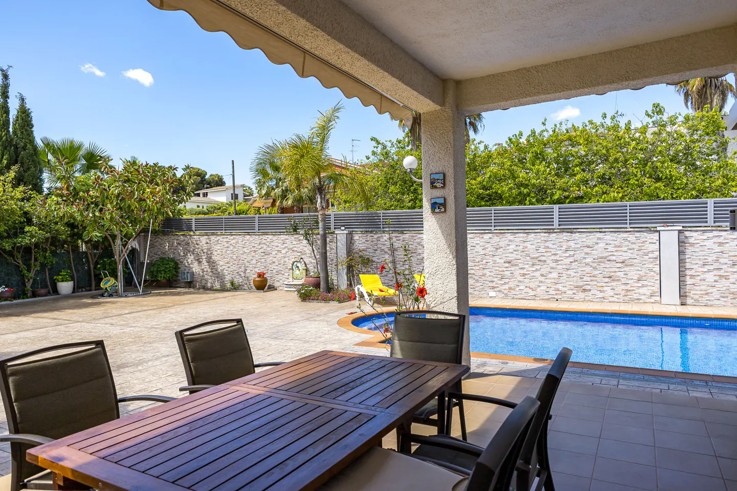 Outdoor patio with a dark wood table and chairs, a blue swimming pool, and lush green trees under a bright blue sky.