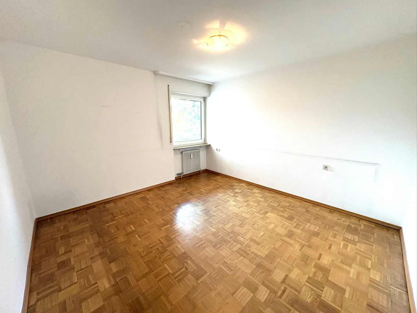 Empty room with parquet floor, white walls, and a window with a radiator below. Ceiling light is on.