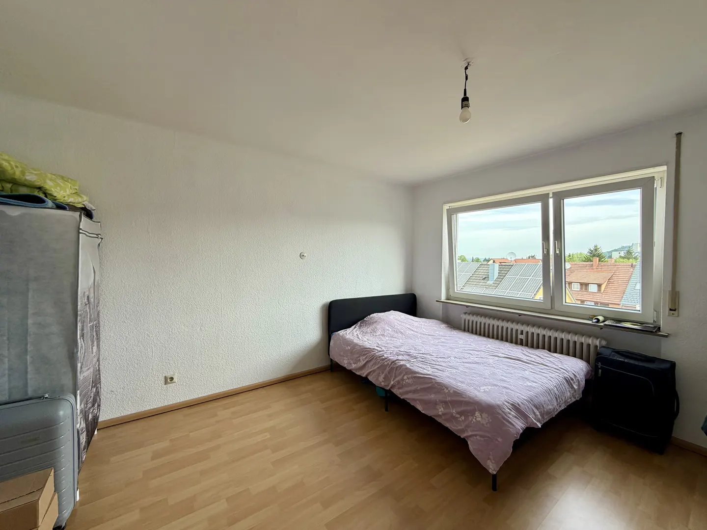 Bedroom with a bed covered in a purple patterned sheet, a window with a view, and a wooden floor.