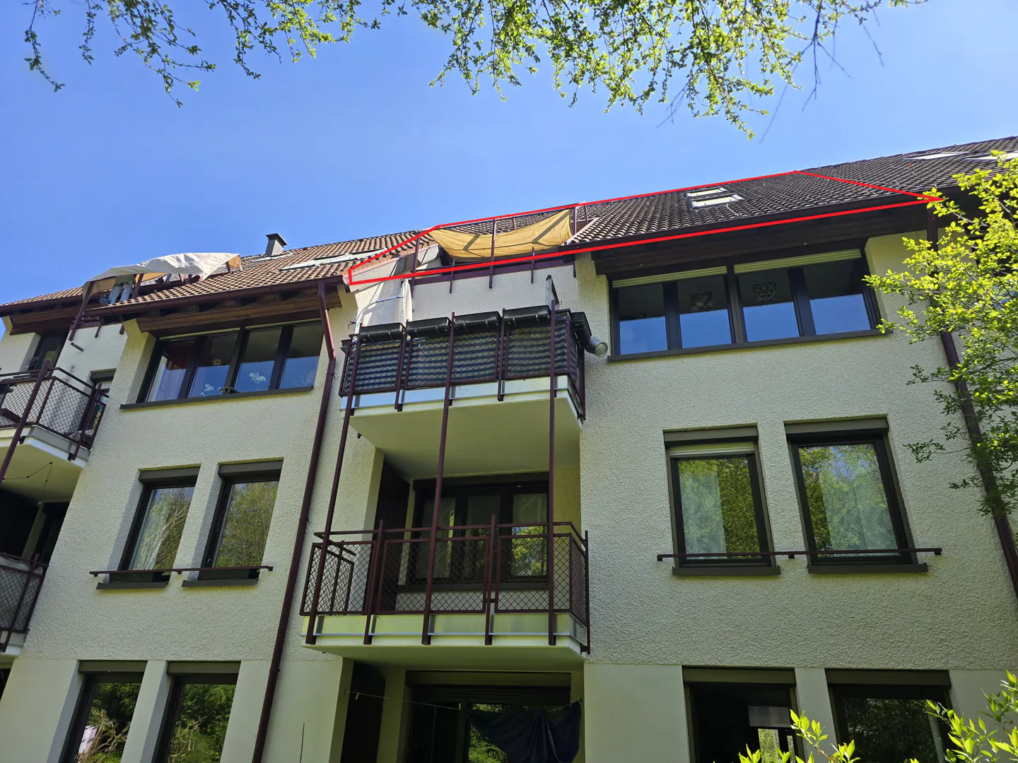 Exterior view of a three-story apartment building with balconies, brown window frames, and a red roofline against a blue sky.