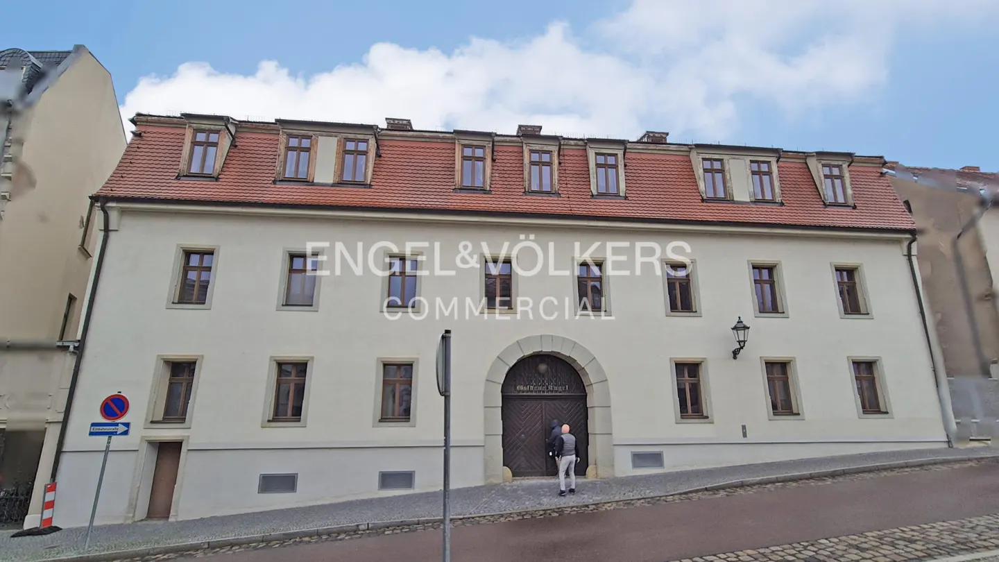 Exterior of a cream-colored commercial building with a red tile roof and many windows. A man stands at the arched wooden doorway. "Engel & Völkers Commercial" is printed on the building.