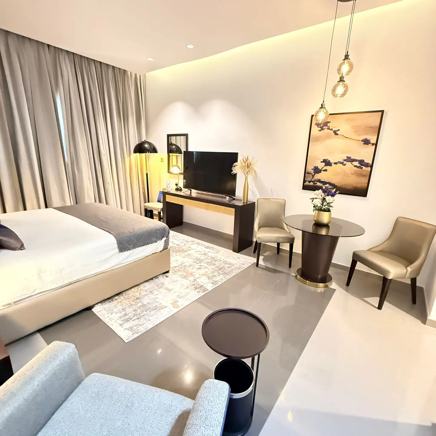 A modern bedroom with a bed, TV, table, chairs, and a painting on the wall. The room is decorated in neutral colors.