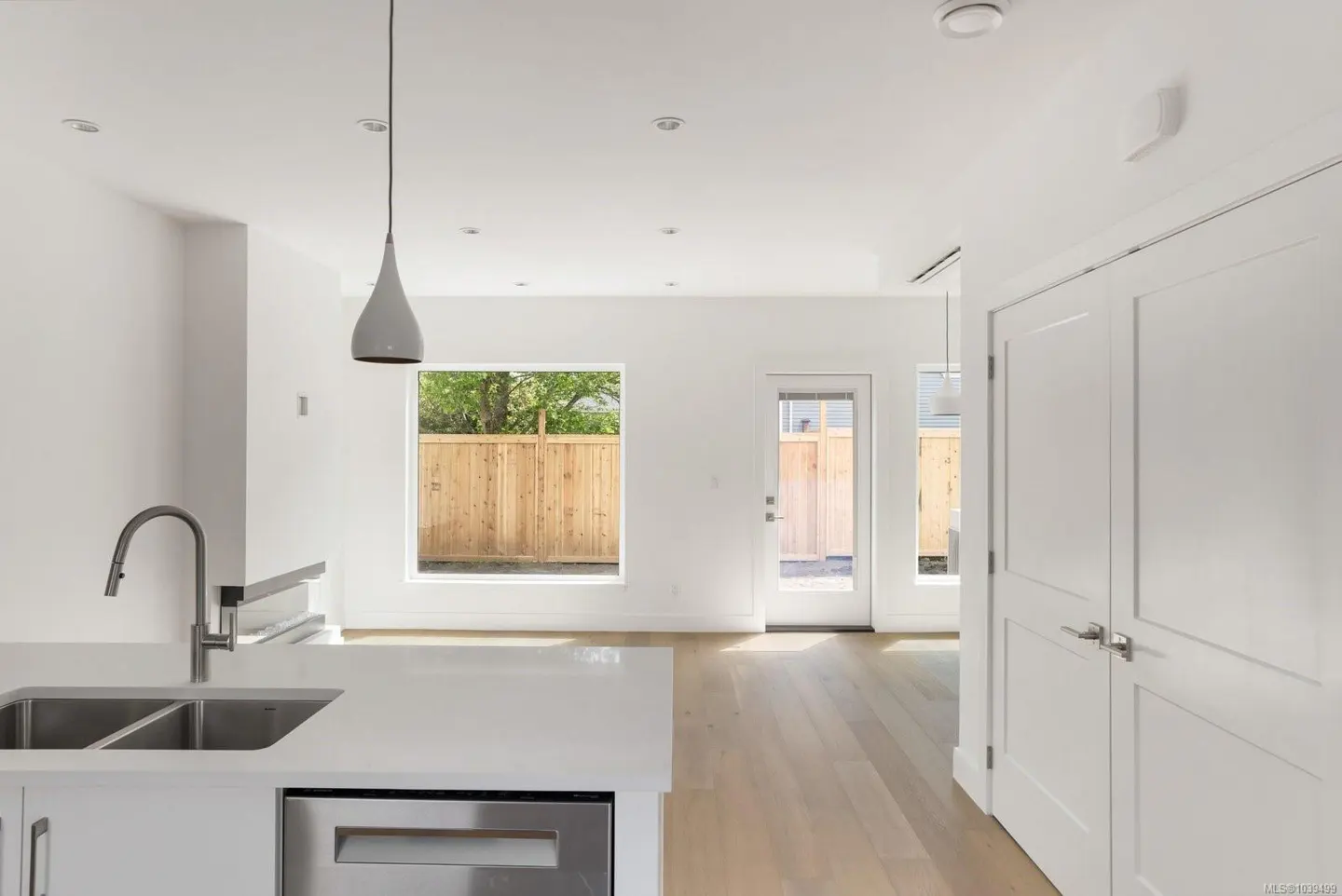 Modern white kitchen with stainless steel sink, light wood floors, and a view of a wooden backyard fence through windows.