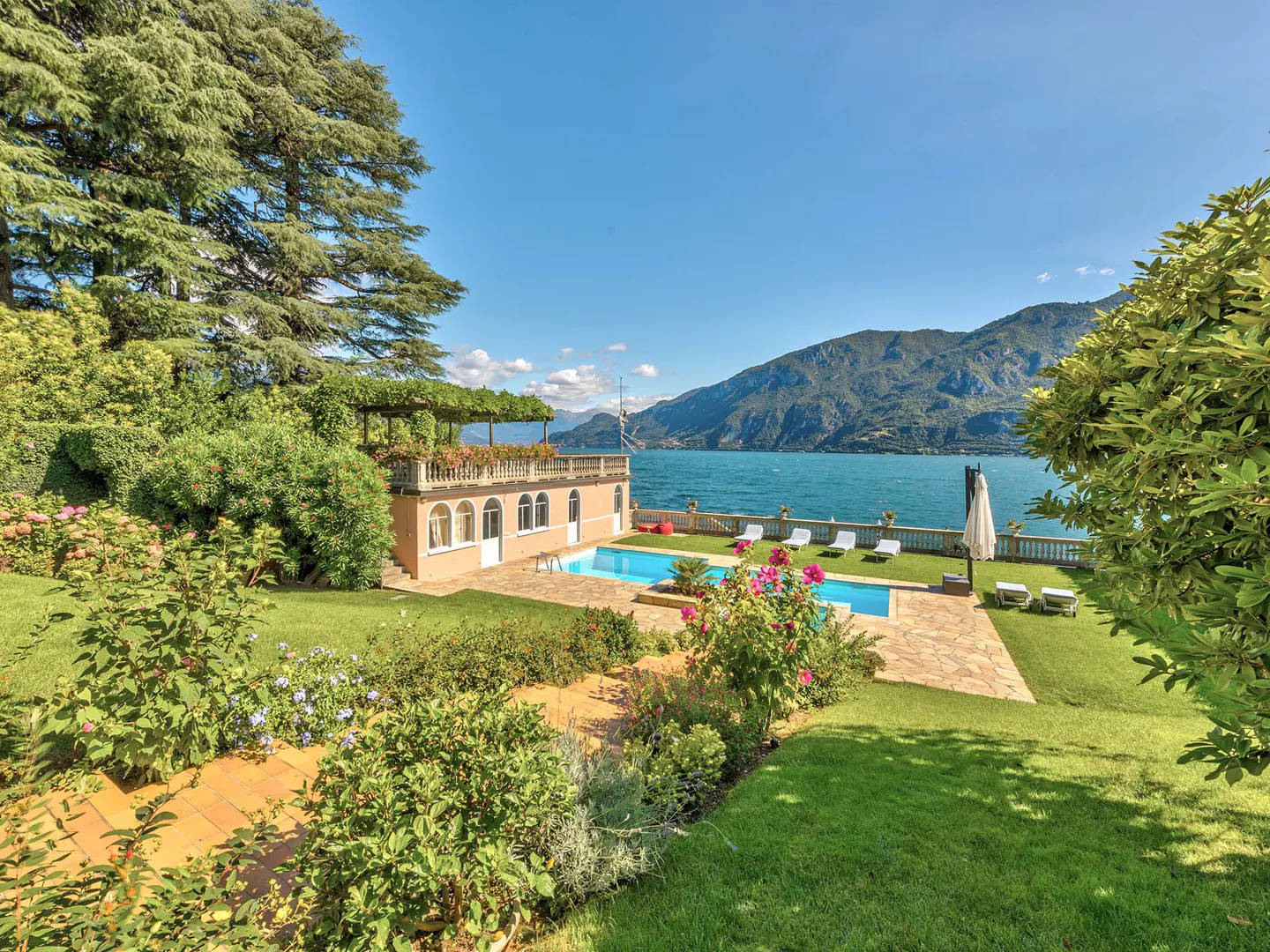 Luxury villa with a pool overlooking a lake and mountains. The villa is peach-colored with a balcony and arched windows. Green lawn and trees surround the property.