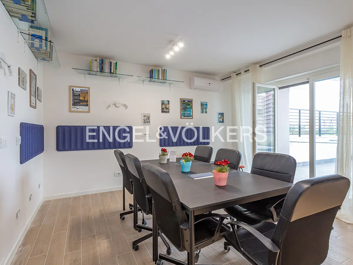 Bright office space with a dark conference table, black chairs, and a balcony with a view. Engel & Völkers logo on the wall.