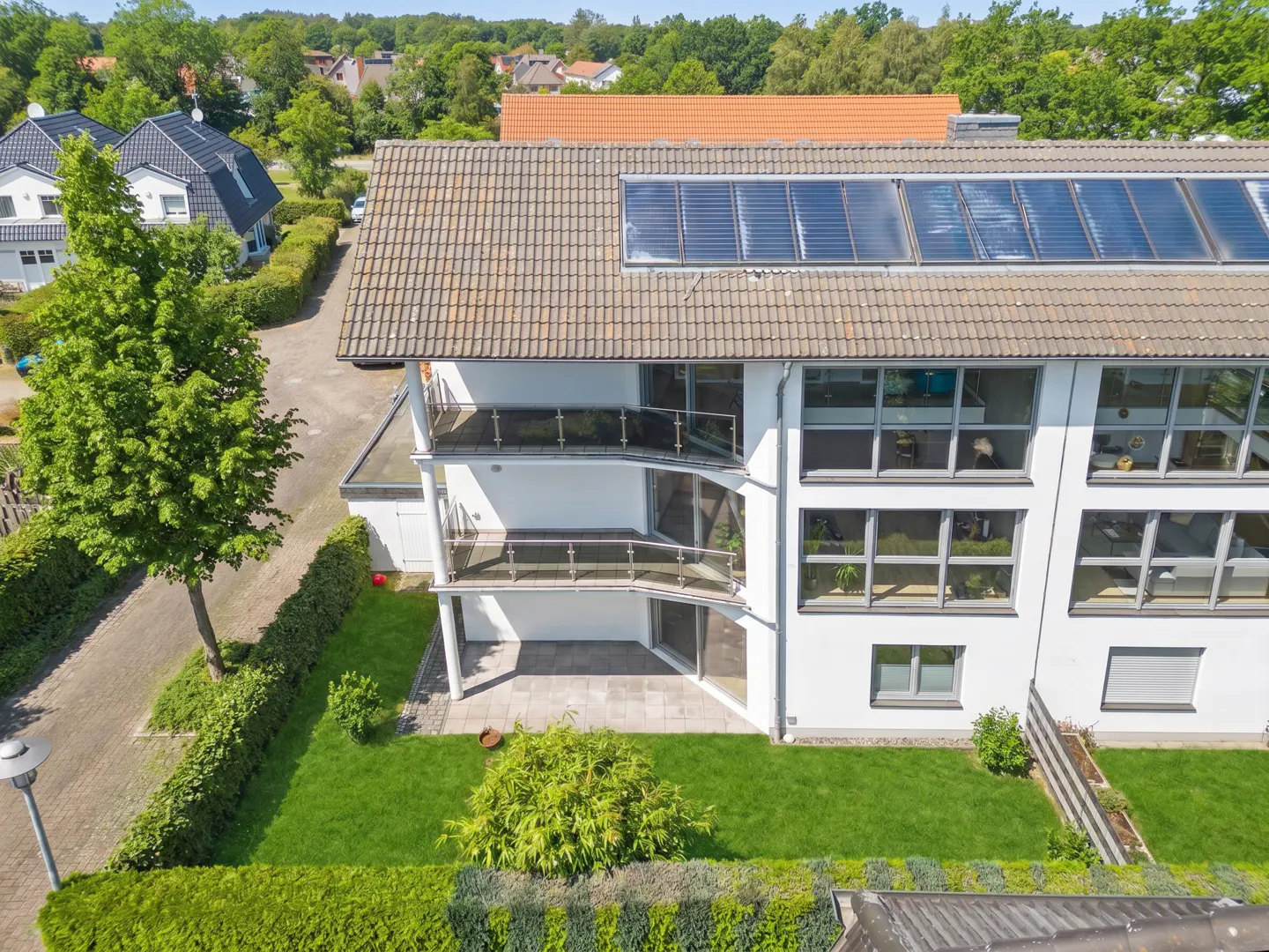 Aerial view of a modern white house with balconies, large windows, and solar panels on a tiled roof, surrounded by green lawns and trees.