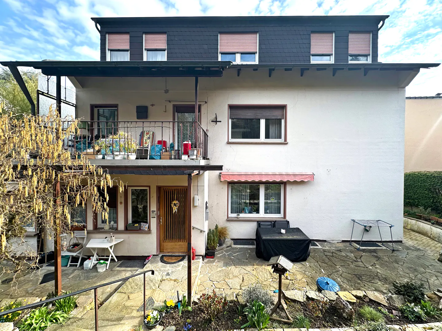 Two-story house with a balcony, brown shutters, and a stone patio. A tree with yellow flowers is on the left.