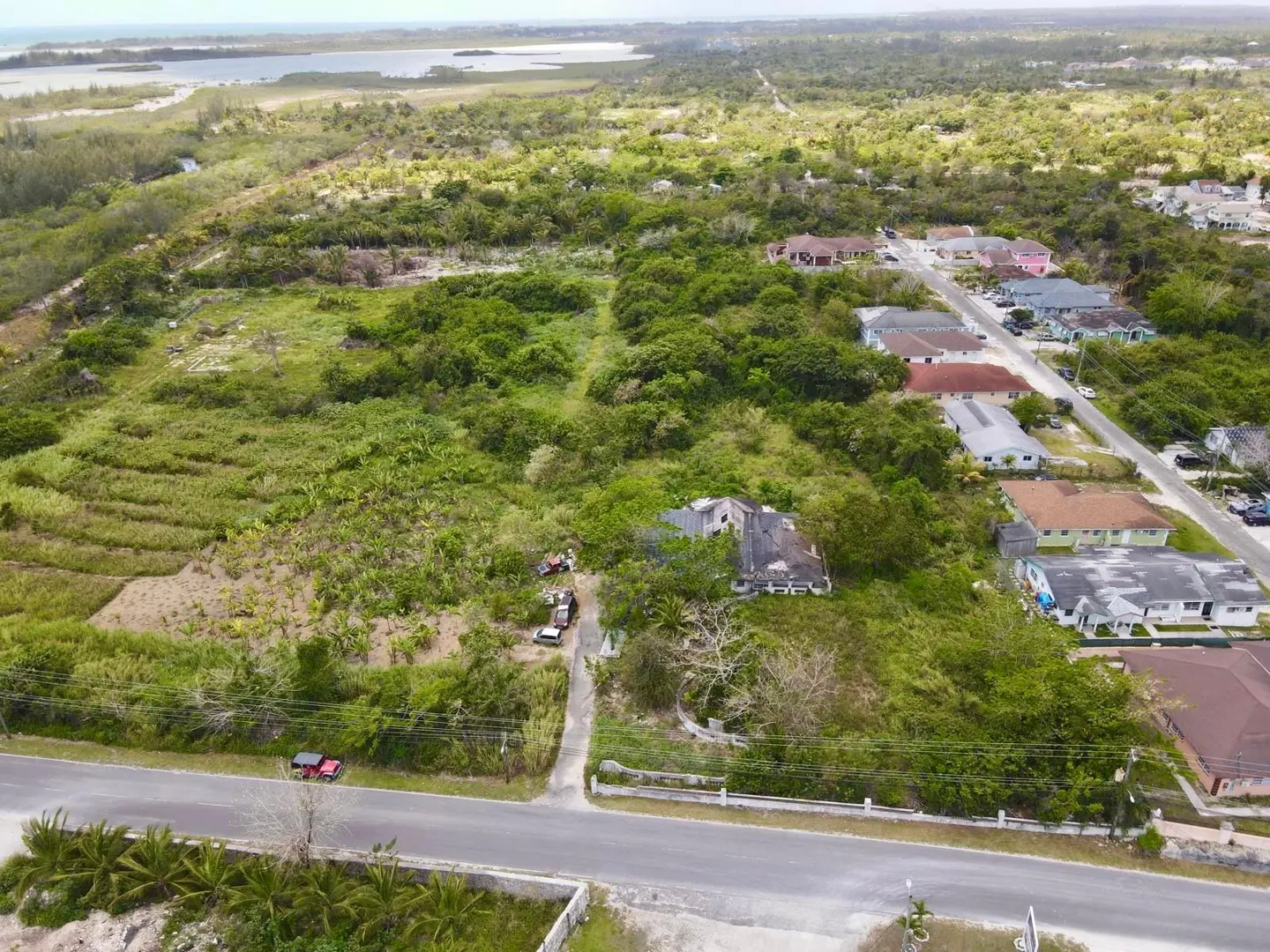 Aerial view of a large, grassy lot with a house, adjacent to a road and residential neighborhood near a coastal inlet.
