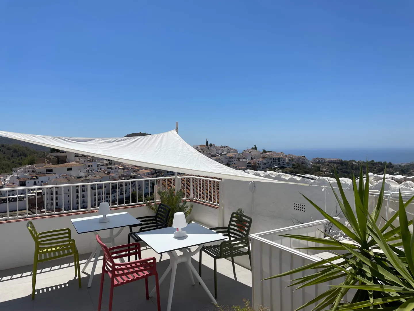 Outdoor patio with colorful chairs and white tables under a white awning, overlooking a white village and the sea.