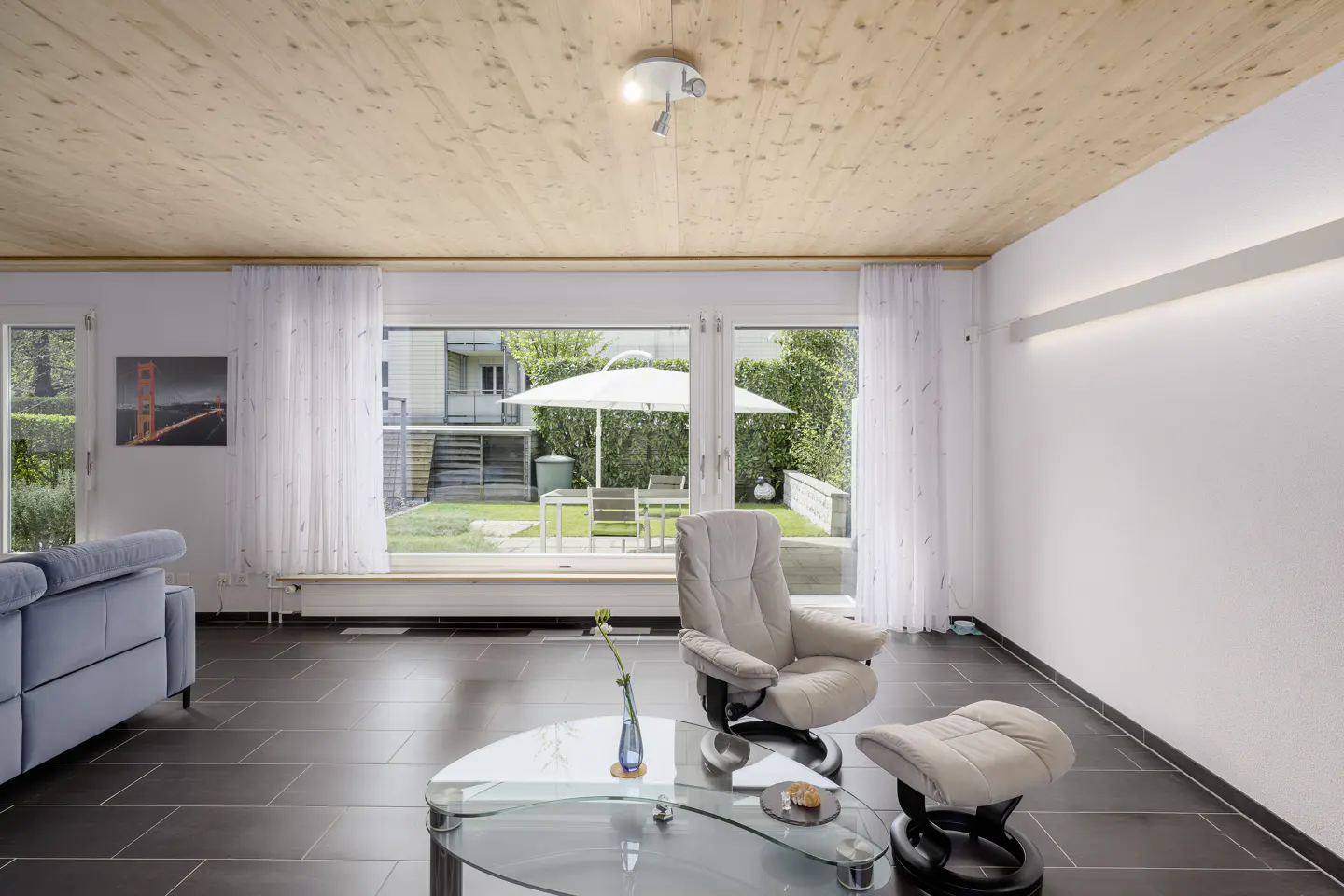 Bright living room with wood ceiling, tile floor, and large windows overlooking a green lawn with patio furniture.