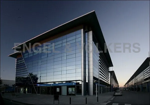 Modern commercial building with glass facade and flat roof, reflecting the sky. Street view with other similar buildings in the background.