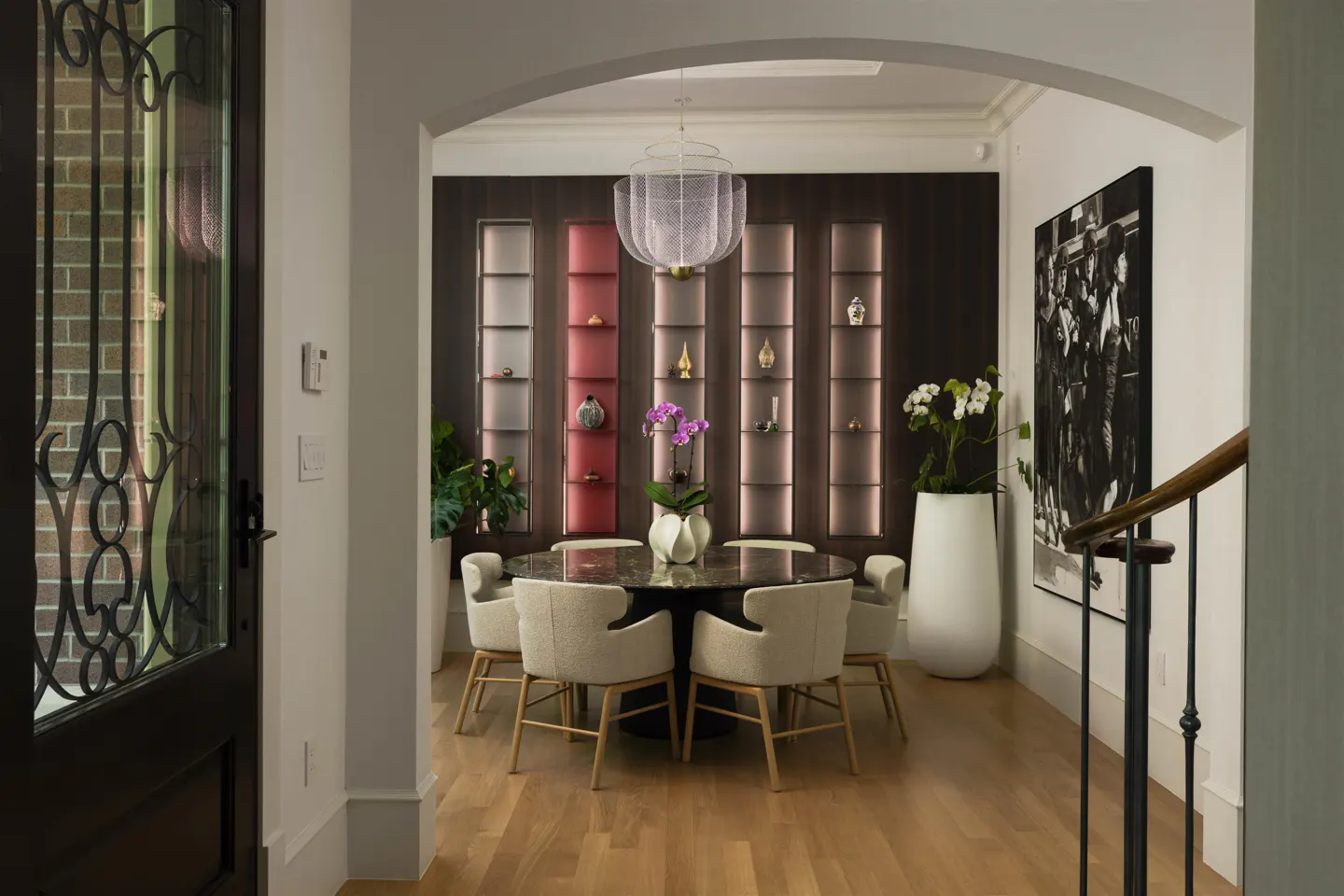 Elegant dining room with a round table, beige chairs, and a decorative chandelier. Built-in shelves display art, and a black and white print hangs nearby.
