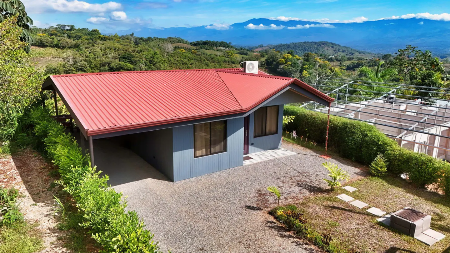Exterior of a blue house with a red roof, carport, gravel driveway, and mountain views.