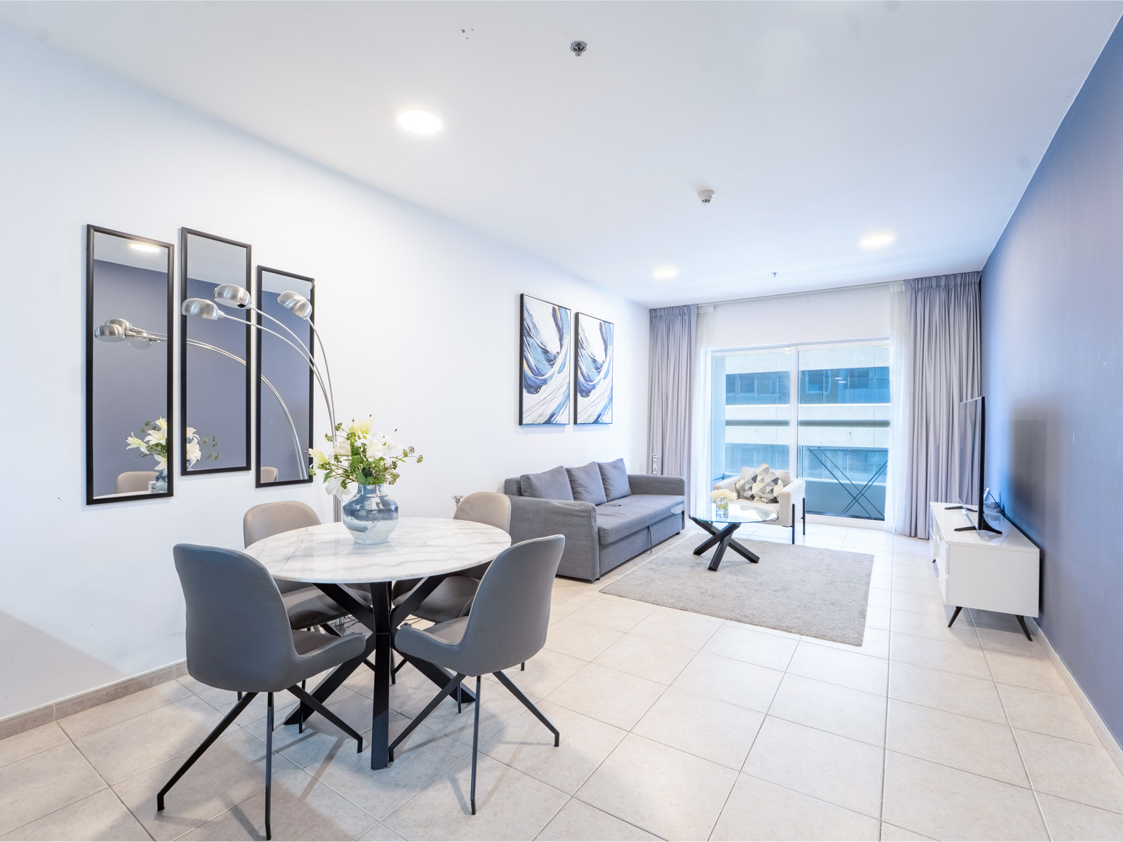 Modern open-plan living room with a marble dining table, grey chairs, sofa, and large windows with light curtains.