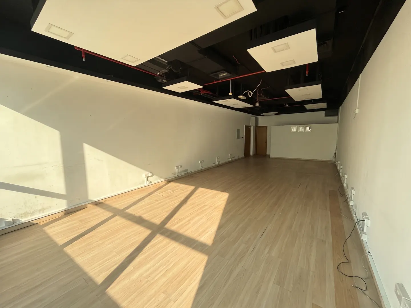 Empty commercial space with light wood floors, white walls, and a black ceiling with square lighting fixtures.