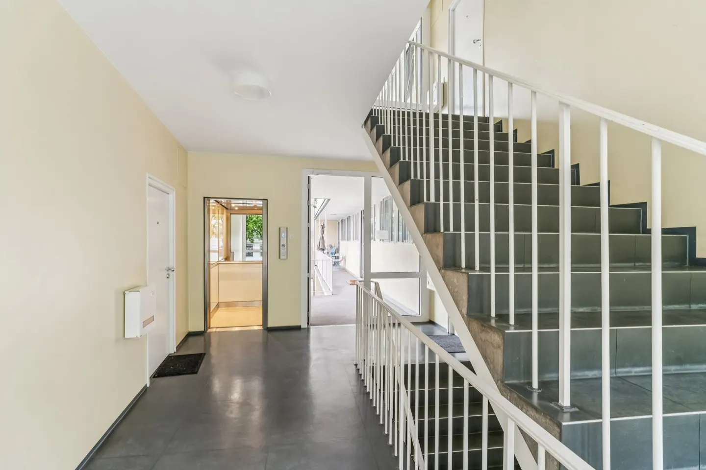 Hallway with gray tile floor, cream walls, and black stairs with white railing. Elevator and doorway visible.