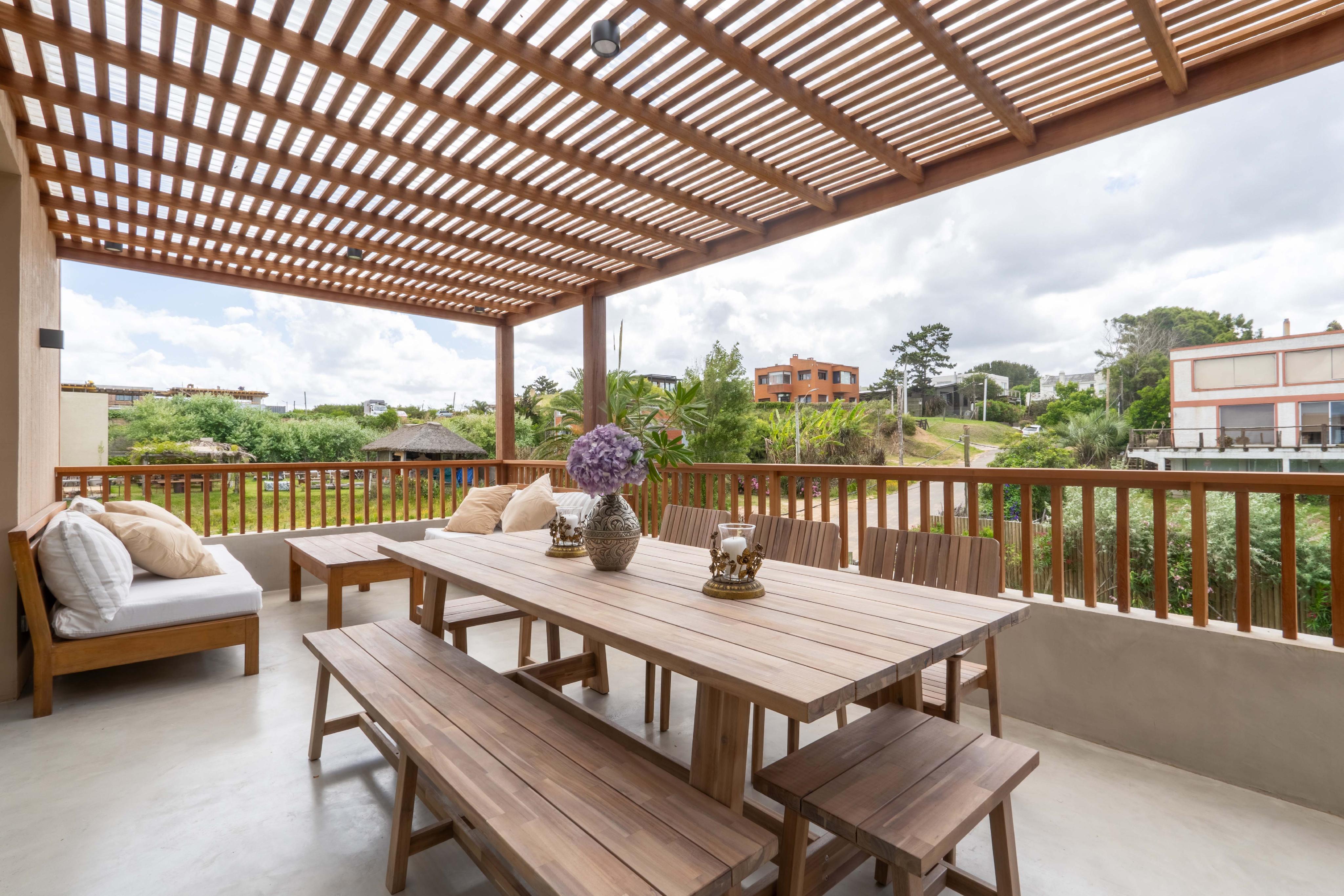 Outdoor patio with wooden pergola, table, benches, and chairs. Pillows on a daybed. View of houses and greenery in the background.