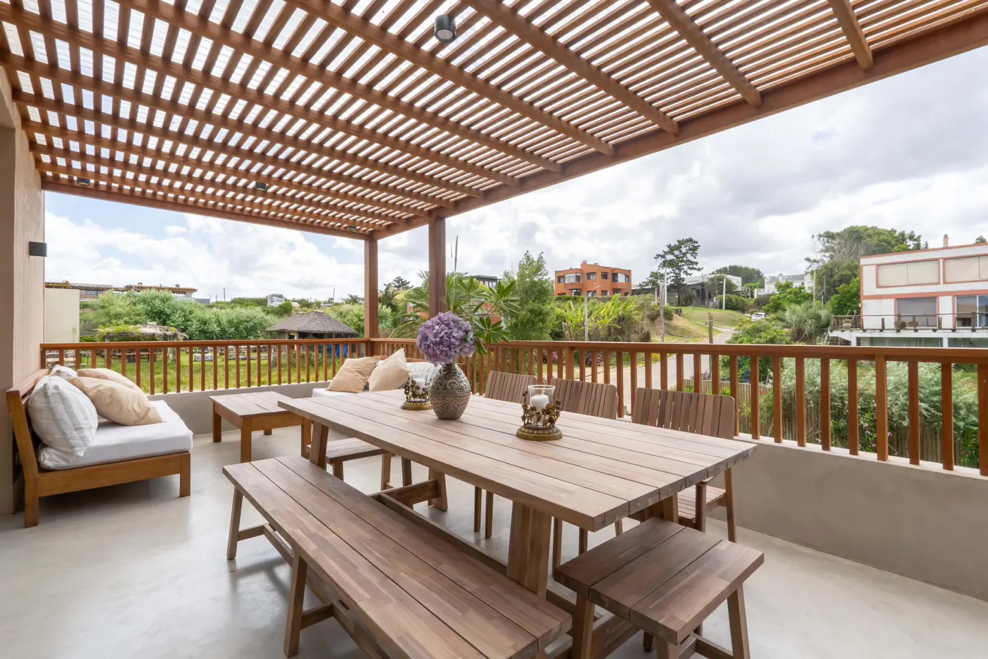 Outdoor patio with wooden pergola, table, benches, and chairs. Pillows on a daybed. View of houses and greenery in the background.