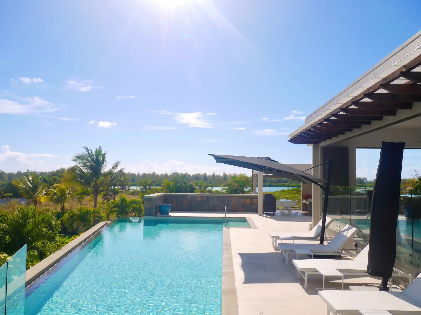 Luxury villa with infinity pool overlooking lush greenery and a blue sky. White lounge chairs sit on the patio.