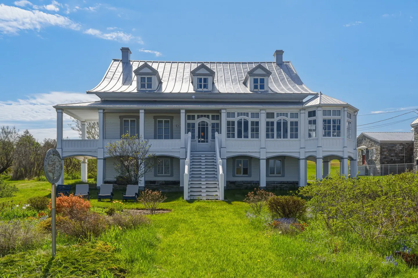 A two-story white house with a metal roof, raised on pillars, with a central staircase and a lush green lawn.