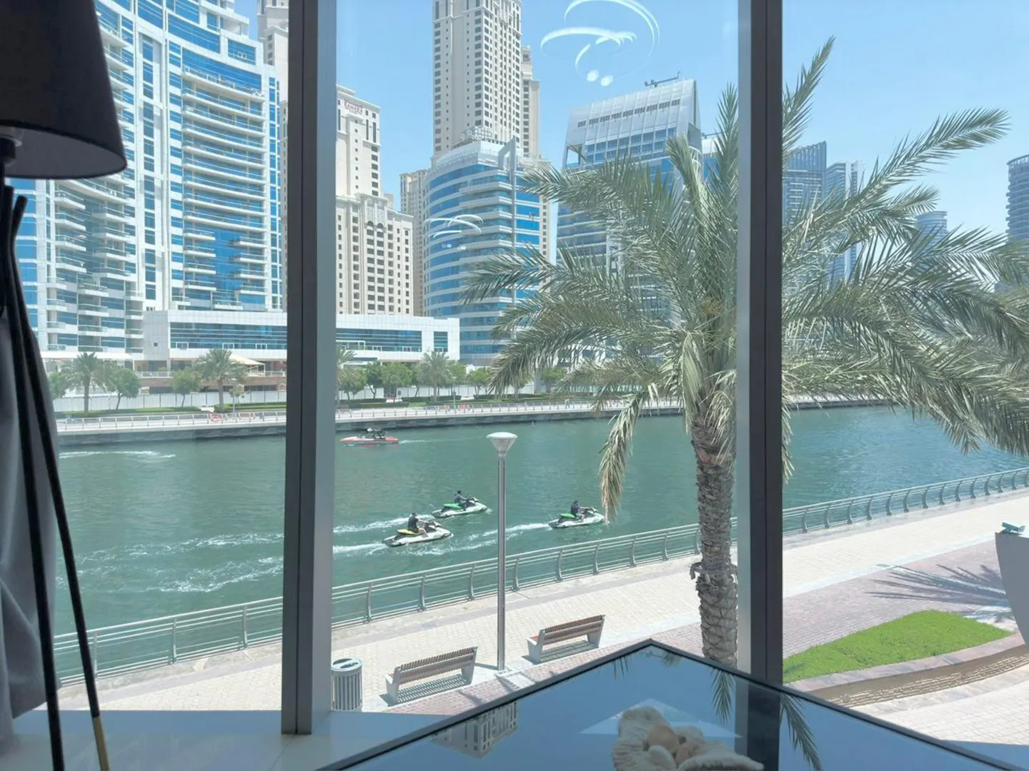 View from a window of a waterfront cityscape with jet skis on the water, palm trees, and modern buildings.