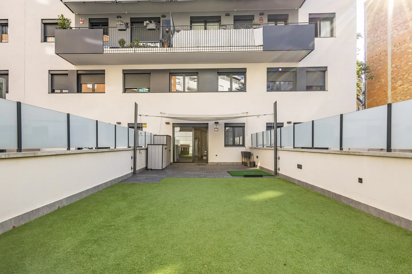 Apartment building with a private outdoor space. Green artificial grass, white walls, and a glass fence. Balconies above.