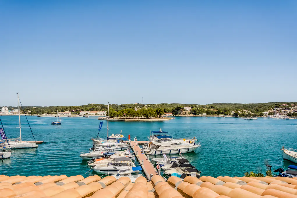 View of a marina with boats docked on turquoise water, seen from a rooftop with orange tiles under a clear blue sky.