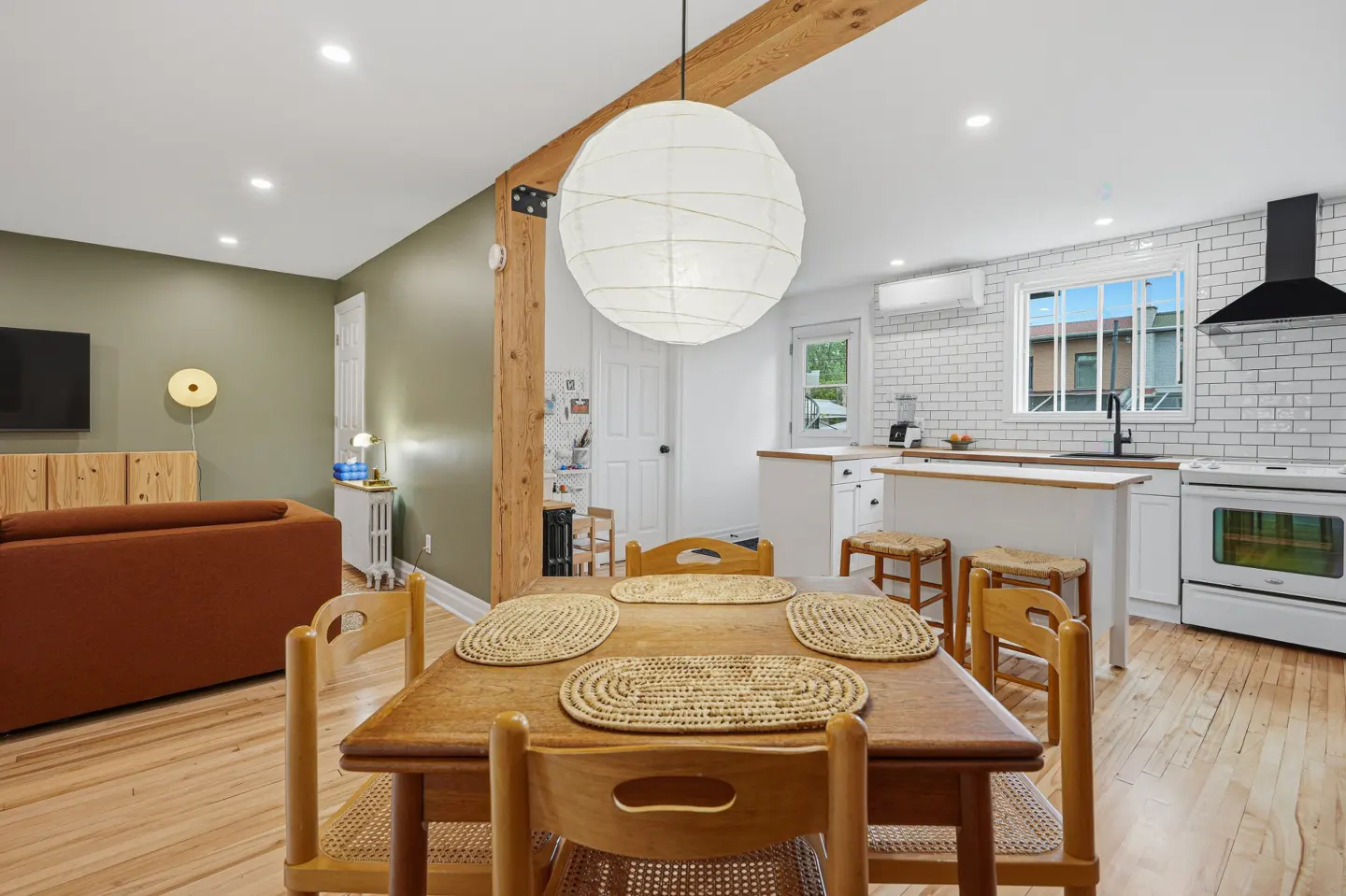 Open-concept living space with a wooden dining table, chairs, and a white paper lantern. The kitchen has white cabinets and subway tile.