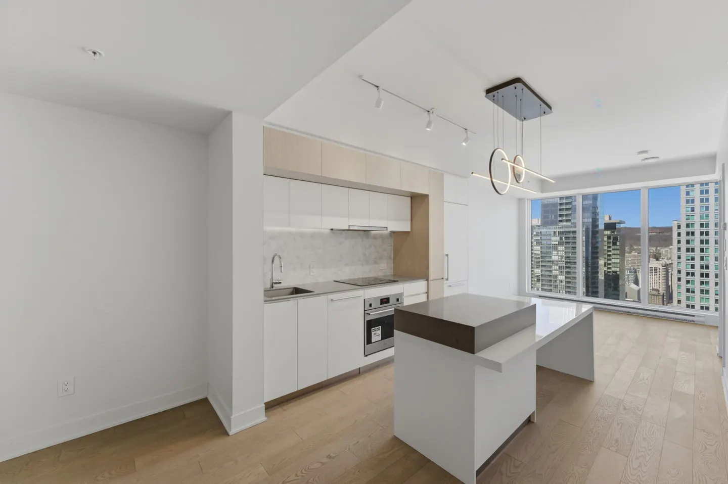 Modern kitchen with white cabinets, island, and stainless steel appliances. Large windows offer a city view. Hardwood floors and stylish lighting.