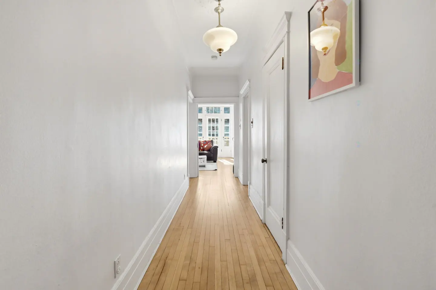 Hallway with light wood floors and white walls. A painting hangs on the right wall. A living room is visible at the end of the hall.