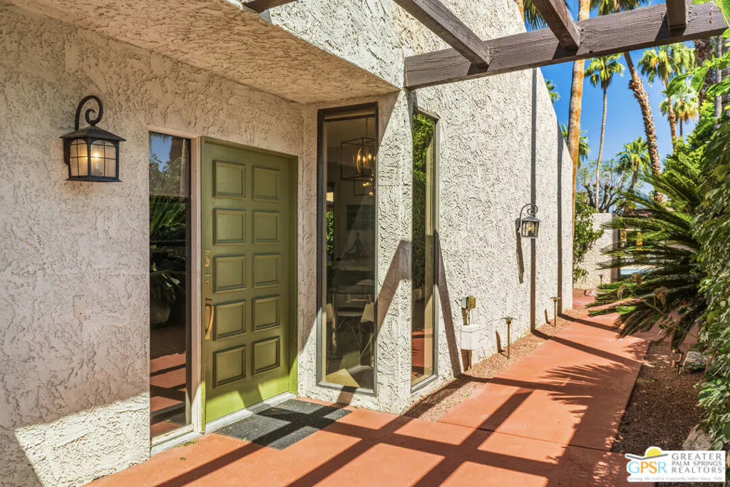 Exterior shot of a stucco house with a green door, black lantern, and a red walkway. Palm trees are visible in the background.
